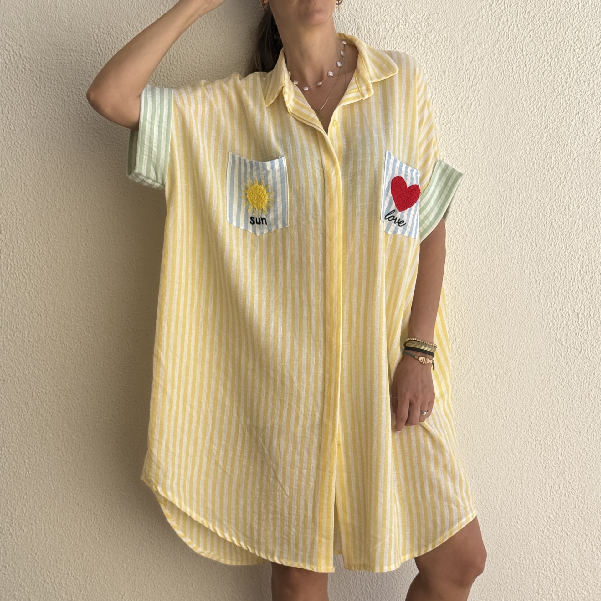 Camisa de riscas amarelas com bolsos bordados e mangas curtas verdes conta parede branca.