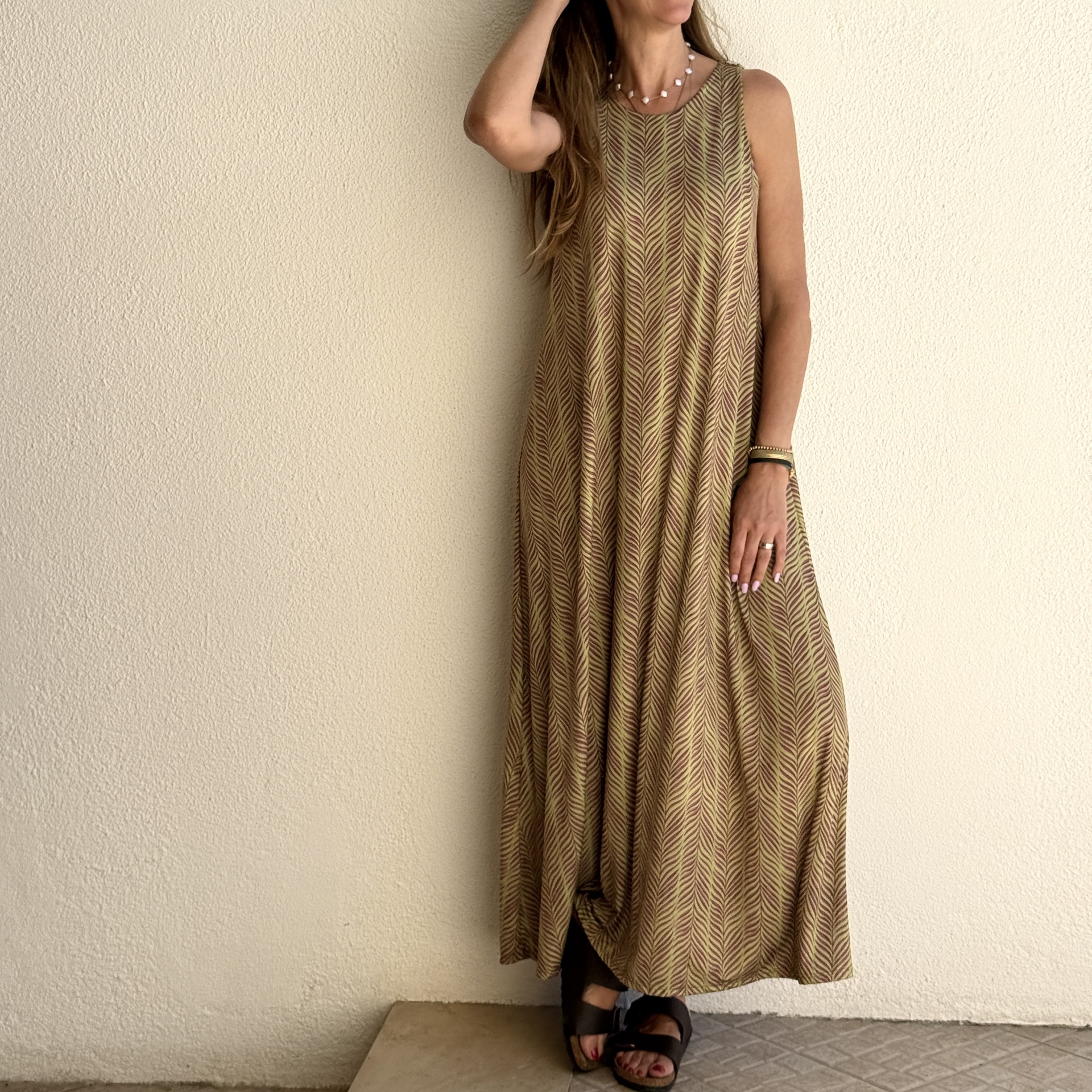 Vestido comprido com padrão de linhas onduladas castanhas e verdes, usado por mulher com colar e sandálias pretas.