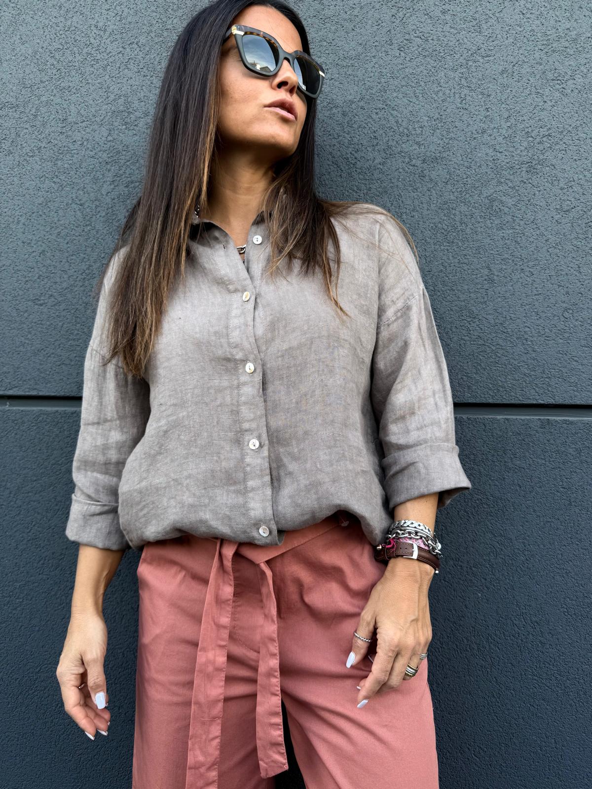 Mulher com camisa cinza de linho e calças rosa com cinto em parede cinza