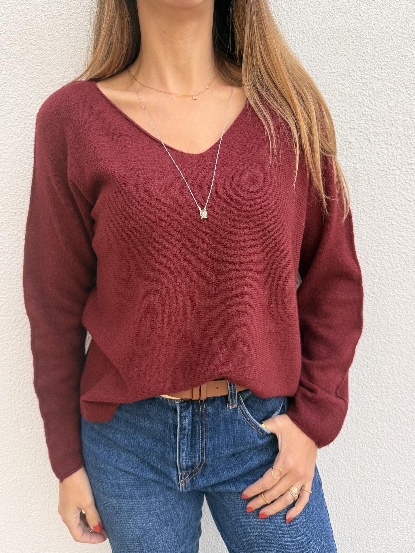 Camisola bordeaux de malha com decote em V usada com jeans azuis e acessórios dourados.
