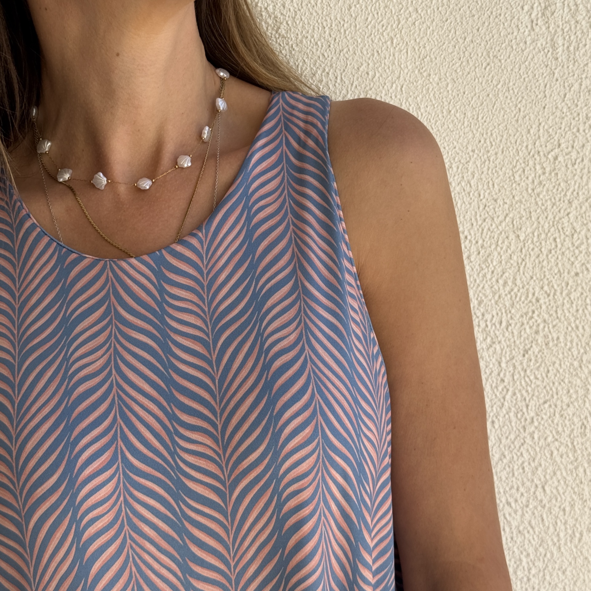 Blusa azul com padrão rosa de folhas, colar de conta branca, parede branca texturada ao fundo