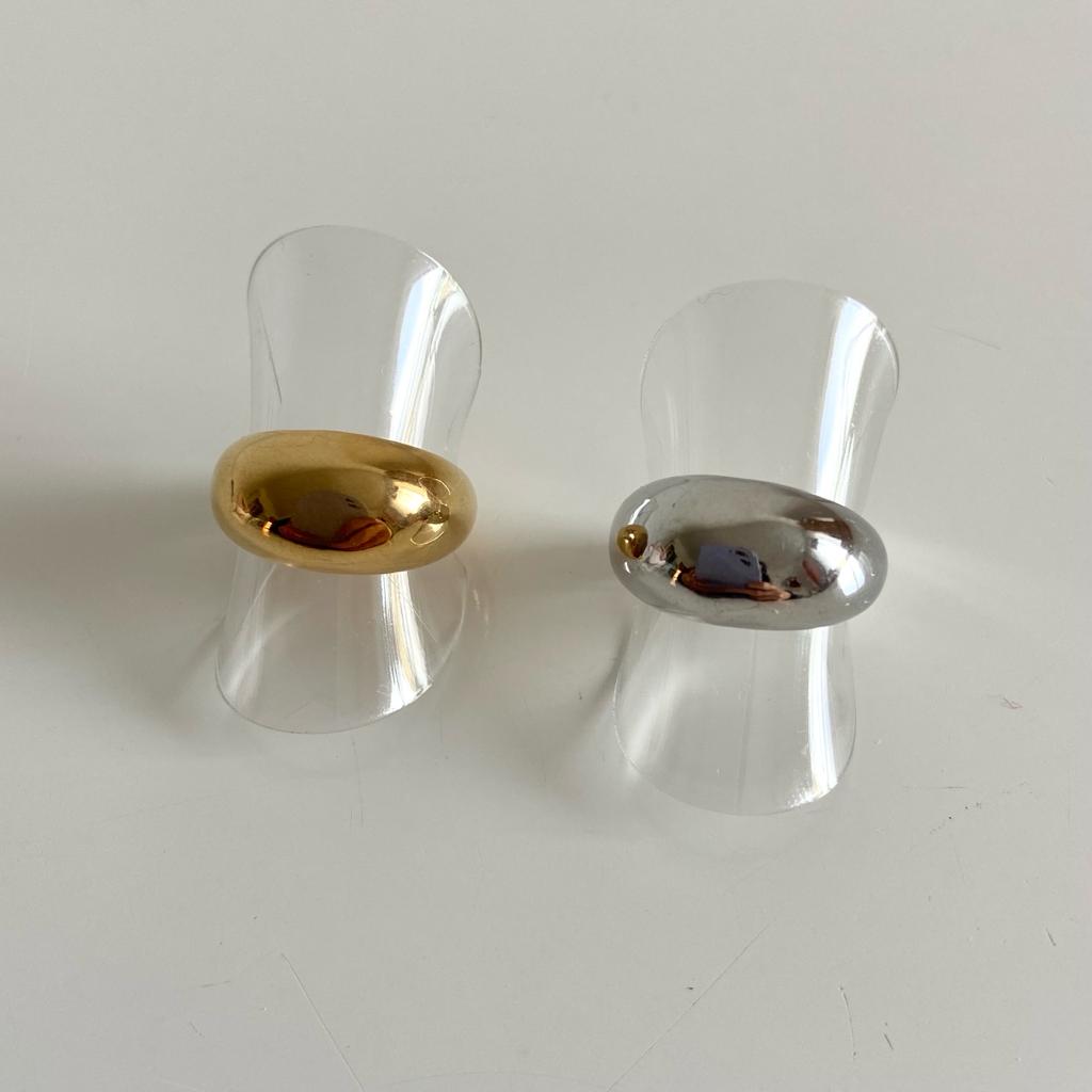 Dois anéis largos, um dourado e outro prateado, em suportes transparentes