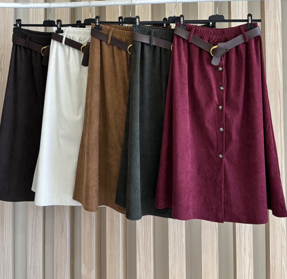 Cinco saias midi de diversas cores em tecido texturizado, penduradas numa barra com cinto castanho e botões frontais na saia vinho
