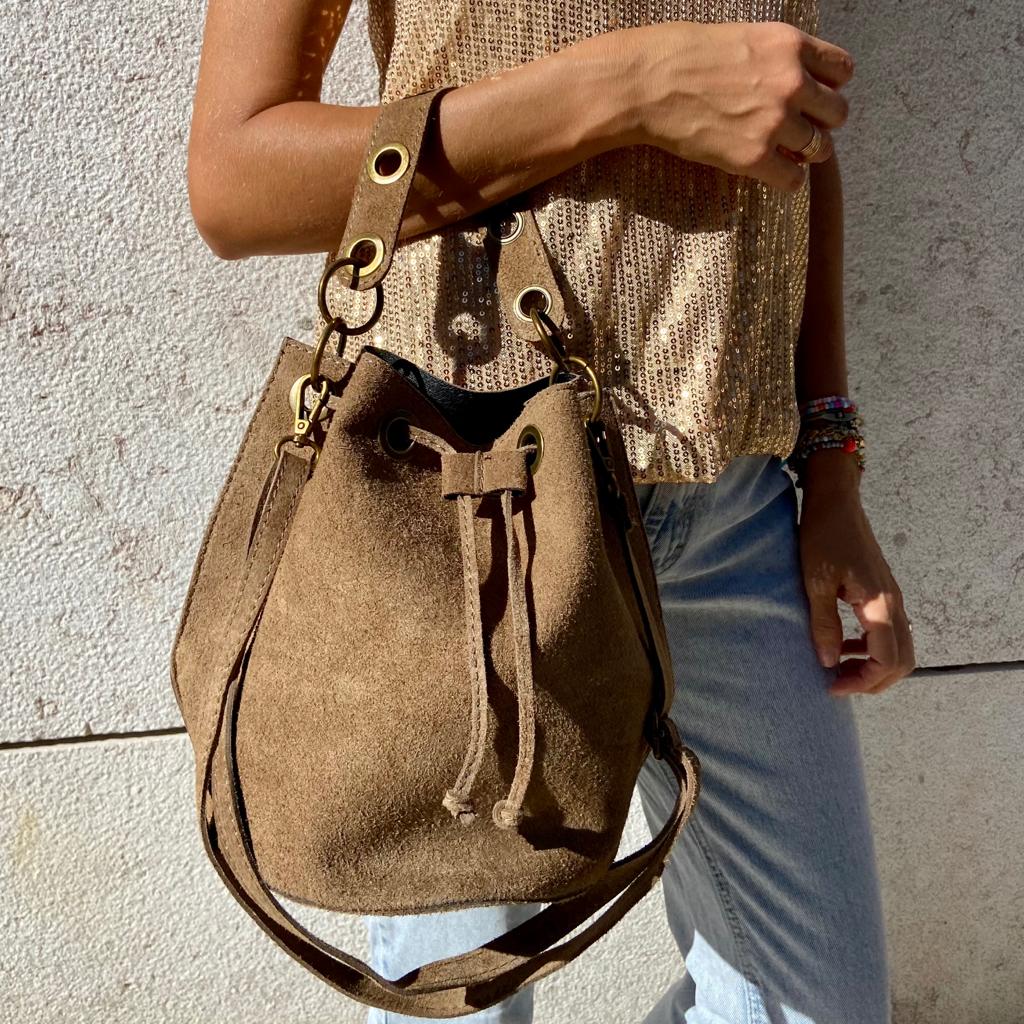 Bolsa de camurça castanha com alças de couro castanho escuro e detalhes dourados, usada por pessoa com camisa bege e jeans claros