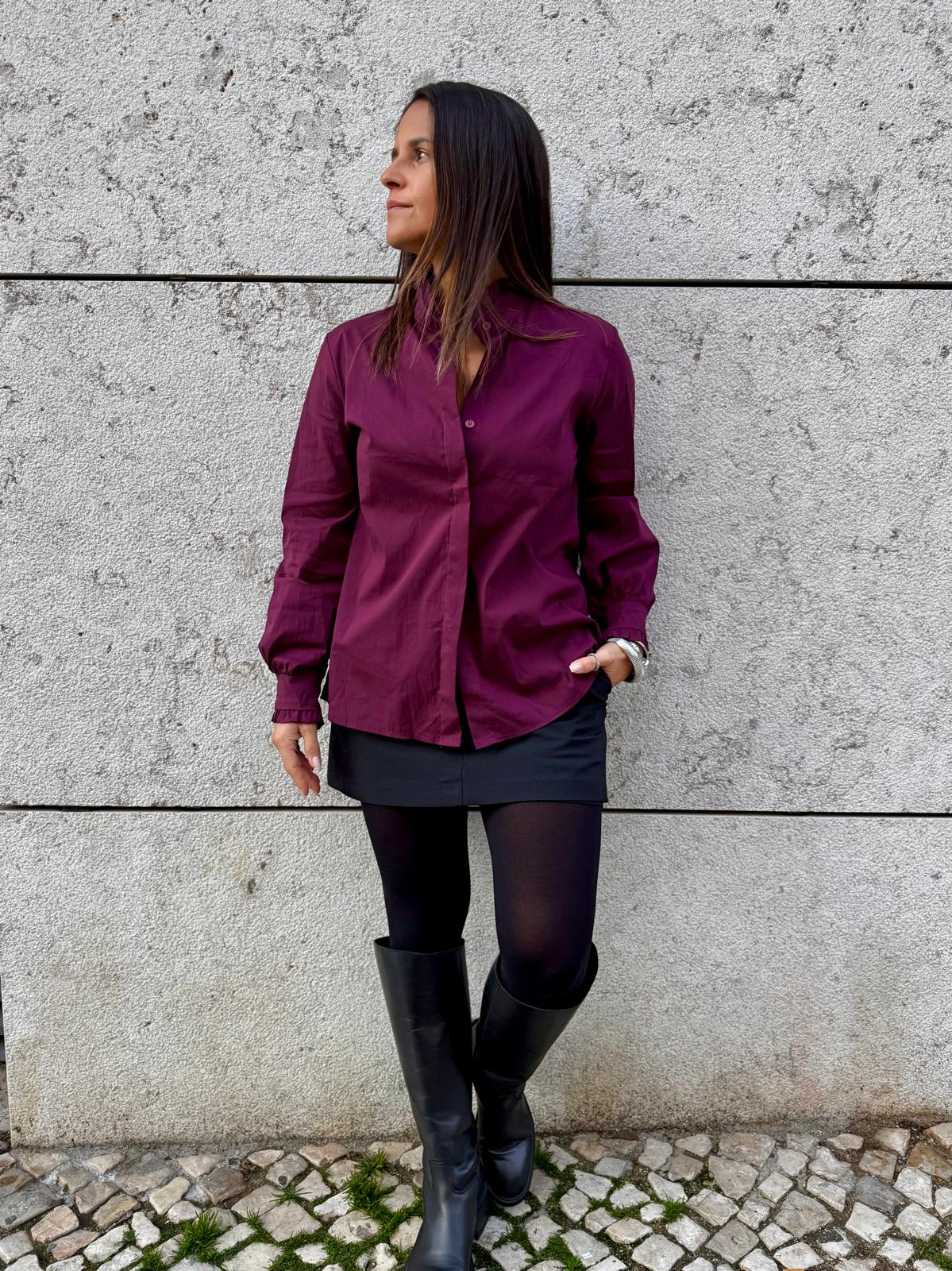 Mulher com camisa vinho, saia preta, meia-calça e botas pretas junto a parede de cimento
