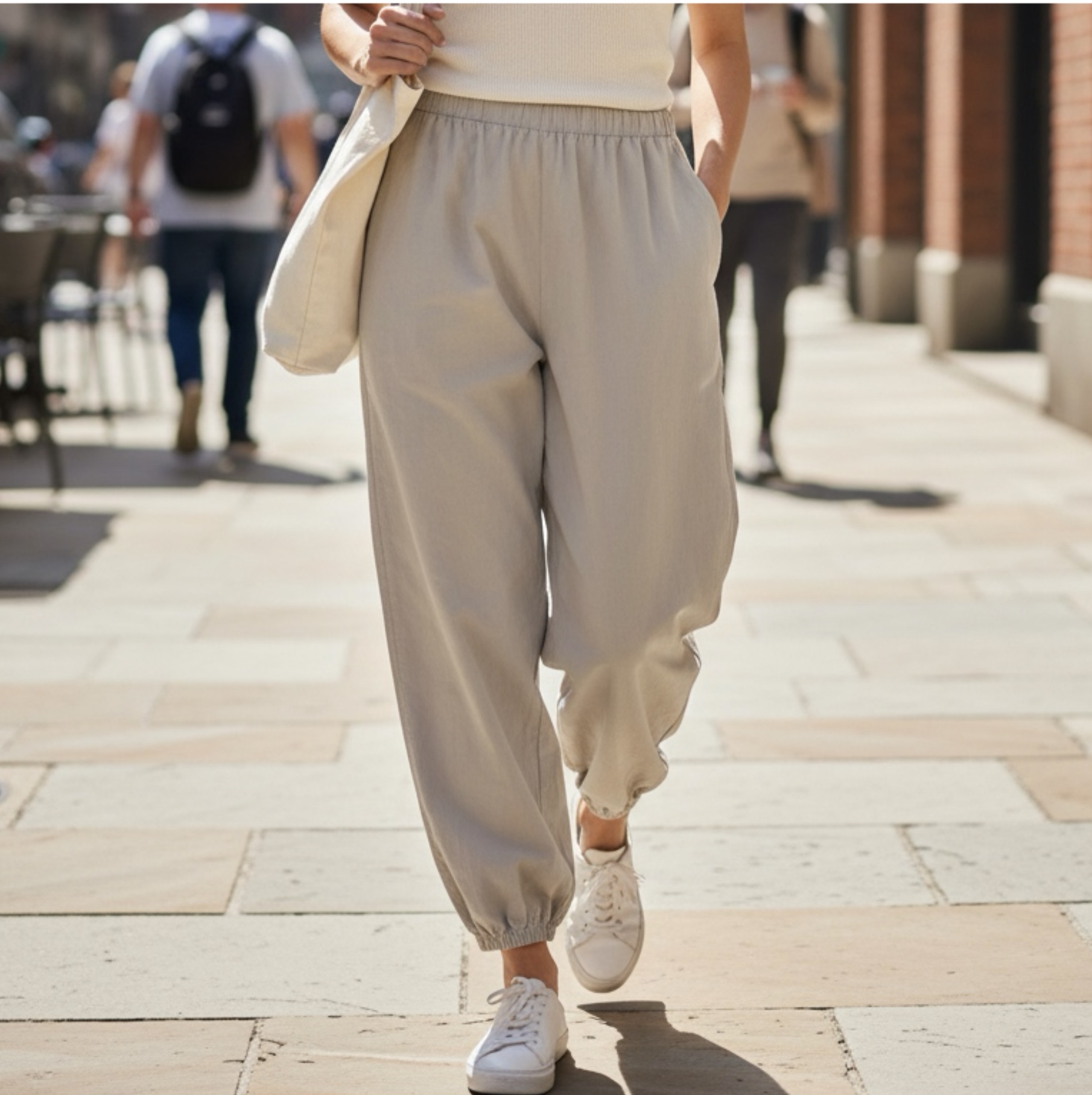 Calças casuais cinza claras com cintura elástica e sapatilhas brancas. Pessoa em rua urbana.