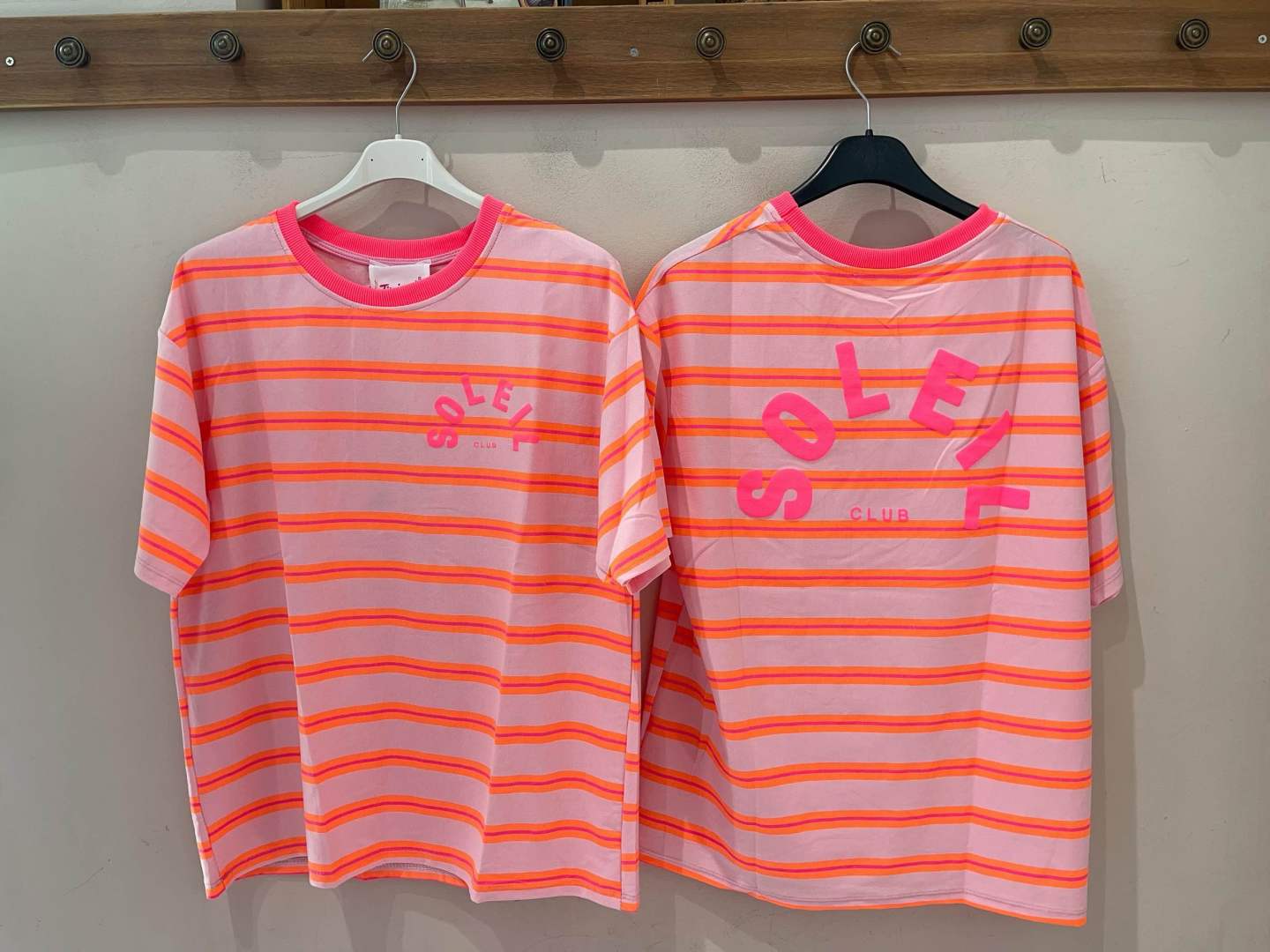 Duas t-shirts listradas brancas e laranja fluorescente com a palavra SOLER em rosa penduradas numa parede.