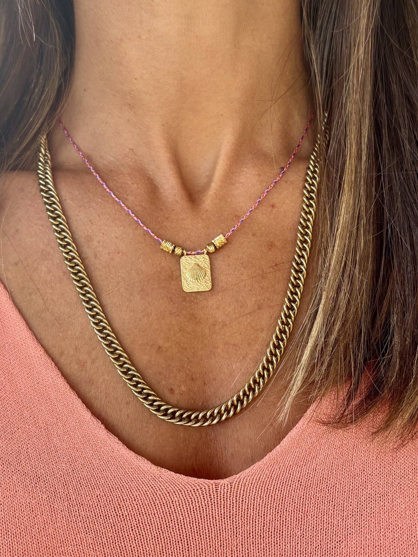 Duas correntes douradas, uma grossa e outra fina com pendente, sobre camisola cor de salmão.