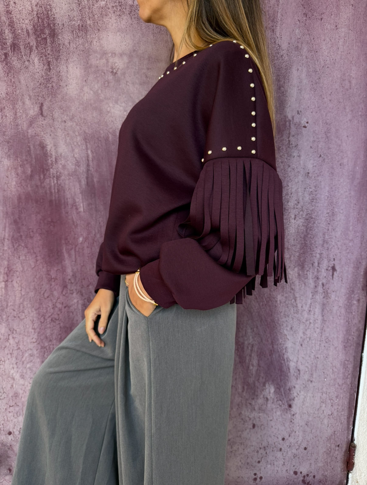 Blusão bordeaux com franjas e tachas, calças largas cinzentas, fundo roxo texturizado