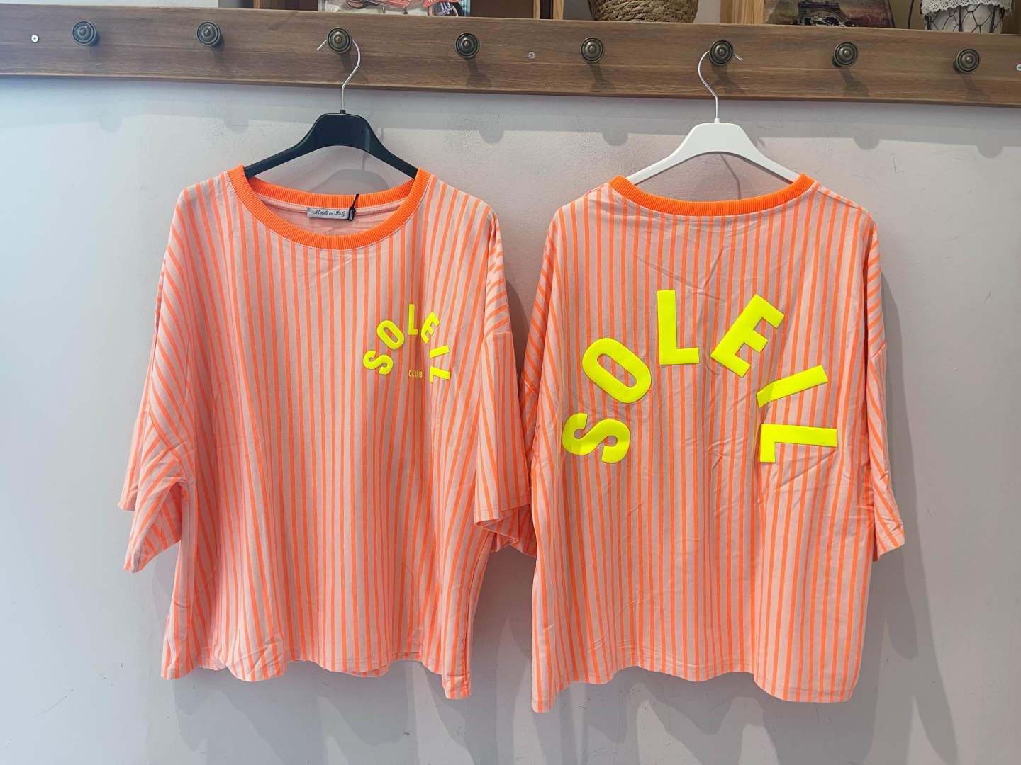 Duas t-shirts riscas laranja e bege com texto amarelo SOLEIL CLUB e SOLEIL, penduradas em cabides.