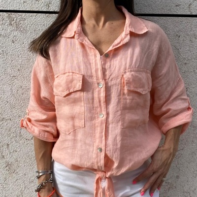 Camisa feminina rosa claro com botões e bolsos, mangas dobradas, amarrada na frente, calças brancas, parede clara ao fundo