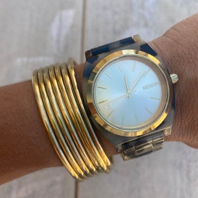 Relógio Nixon dourado com bracelete colorida e pulseiras metálicas douradas no pulso