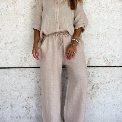 Conjunto feminino bege claro com camisa de manga comprida e calças largas, em fundo de parede de pedra clara
