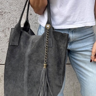 Mala de mão cinzenta em pele camurça com tassel, alças duplas, junto a pessoa com calças de ganga e t-shirt branca.