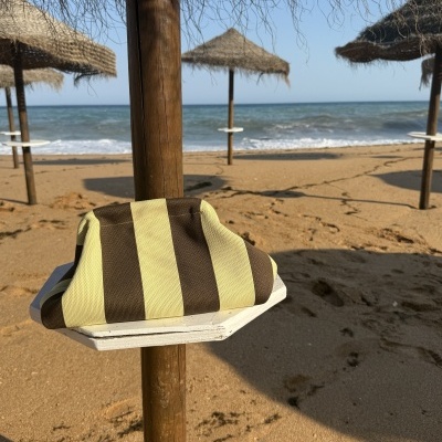 Guarda-sol de palha na praia com toalha às riscas castanhas e amarelas, mar ao fundo e areia dourada.