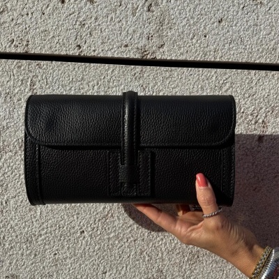 Carteira preta tipo clutch em pele com fecho, sobre fundo de parede clara, segurada por mão feminina