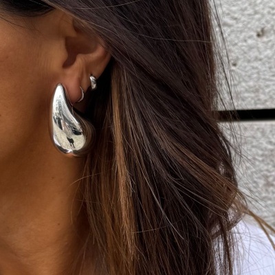 Orelha com dois brincos prateados, cabelo castanho com reflexos loiros, fundo cinza texturizado