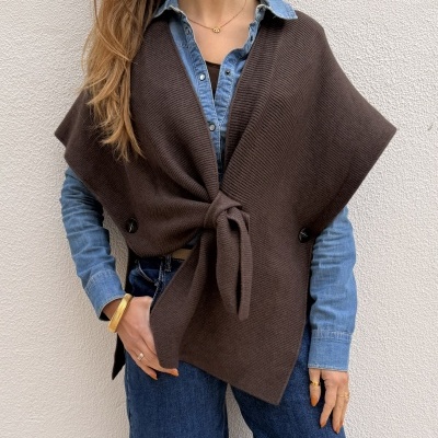 Mulher com colete de malha castanho, camisa de ganga azul e jeans azuis contra parede branca