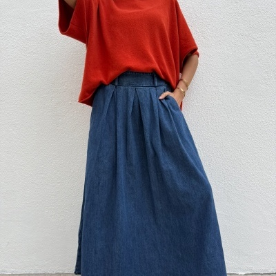 Mulher com camisola vermelha e saia comprida azul escura de denim junto a parede branca