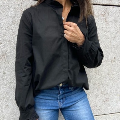 Blusa preta com gola alta e punhos com folhos, usada com jeans azuis, em fundo de parede cinzenta.