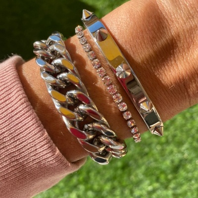 Três pulseiras prateadas no pulso, uma com elos grossos, outra com pedras, e outra com tachas e parafusos, com fundo de relva.