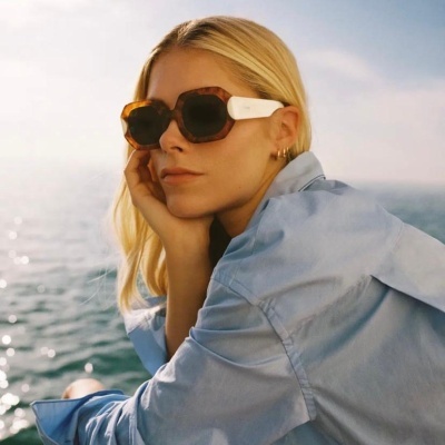 Mulher loira com óculos de sol castanhos e brancos junto ao mar vestindo camisa azul clara