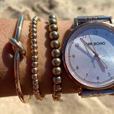 Pulso com relógio MR.BOHO e três pulseiras douradas sobre fundo desfocado. 