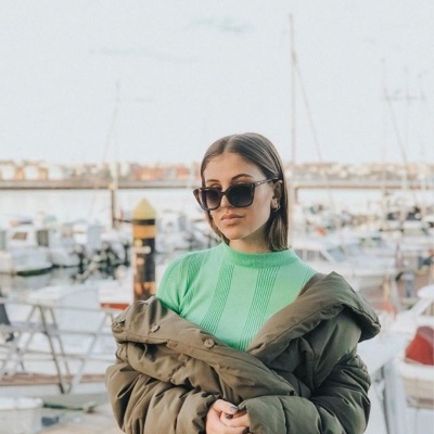 Mulher com camisola verde e casaco acolchoado junto a um cais com barcos