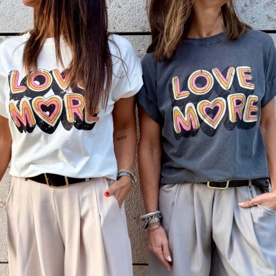 Duas mulheres vestem t-shirts 'LOVE MORE', uma branca e outra cinza, com calças bege contra parede cinza.