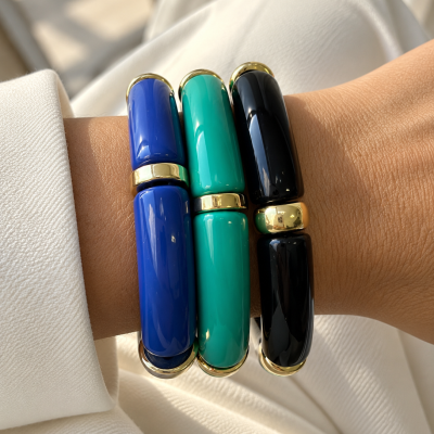 Três pulseiras brilhantes azul, verde e preto com detalhes dourados no pulso de uma pessoa.