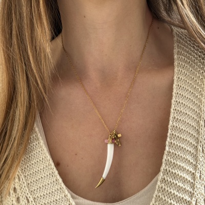 Colar dourado com pendente de dente branco e pingentes, em pescoço de pessoa com roupa bege.