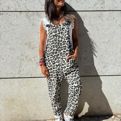 Mulher com macacão estampado animal em tecido, t-shirt branca e sapatilhas brancas, parede de pedra ao fundo.