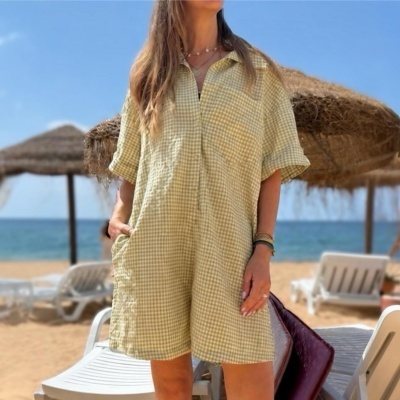 Pessoa com macacão curto amarelo com padrão de quadrados pequenos, ao ar livre na praia com chapéus de palha atrás.