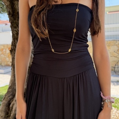 Mulher com vestido preto e colares dourados, pulseiras coloridas, em ambiente exterior com árvore ao fundo.