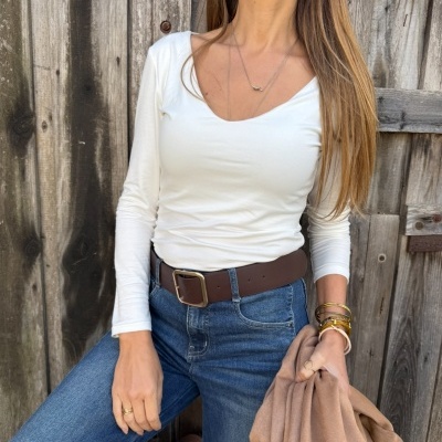 Mulher com camisola branca, jeans azuis, cinto castanho e casaco bege contra porta de madeira