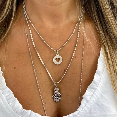 Colares prateados com pendentes de coração e mão Hamsá numa pele clara com blusa branca.