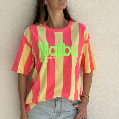 T-shirt riscas amarelas e rosas com texto verde MALIBU usada por mulher com calças de ganga