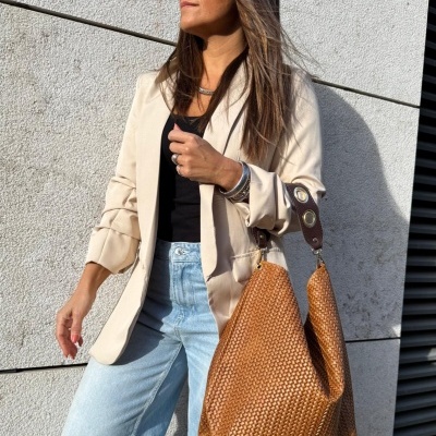 Mulher com casaco bege, top preto, calças de ganga azuis e mala castanha entrelaçada ao ar livre