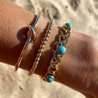 Pulseiras douradas com pedras turquesa num pulso sobre fundo de areia