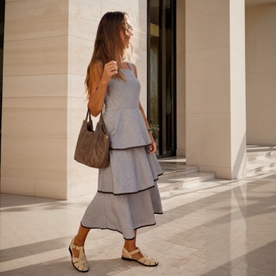 Mulher com vestido azul claro e mala castanha a caminhar num espaço exterior com colunas.