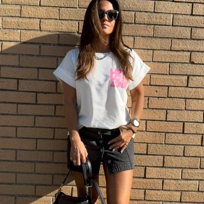 Mulher com blusa branca com estampado rosa e saia preta com botões, óculos escuros e bolsa preta em frente a parede de tijolos