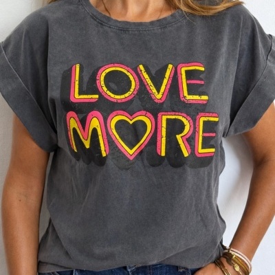 T-shirt cinza escuro com estampa LOVE MORE em amarelo e rosa e coração, uma pessoa veste jeans azul e pulseiras no pulso direito