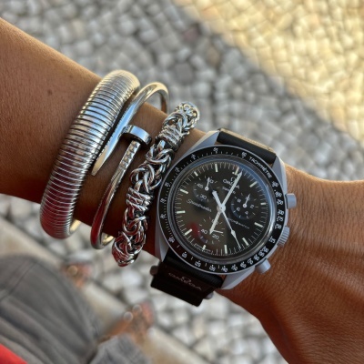 Relógio Omega Speedmaster com pulseiras metálicas prateadas num pulso sobre calçada portuguesa