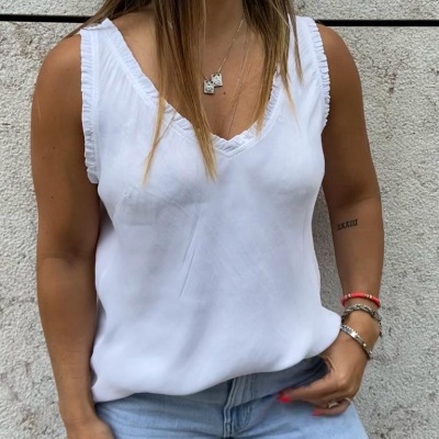 Top branco feminino com decote em V e babados, calças de ganga azuis claras, parede de pedra clara ao fundo.