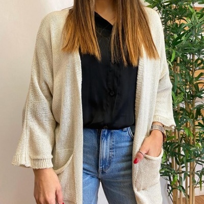 Mulher veste casaco aberto bege de malha com bolsos, camisa preta e jeans azul claro junto a planta em vaso