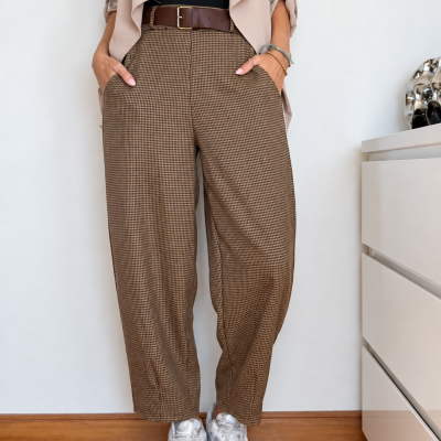 Calças xadrez castanhas com cinto castanho, t-shirt preta, blazer bege e sapatilhas brancas num ambiente interior