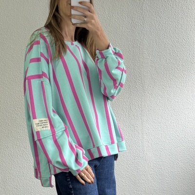 Sweatshirt larga com riscas rosa e azul turquesa e etiqueta no braço junto a parede branca.