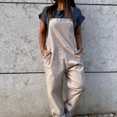 Mulher veste macacão bege com t-shirt cinza e sapatilhas brancas encostada a parede de cimento.