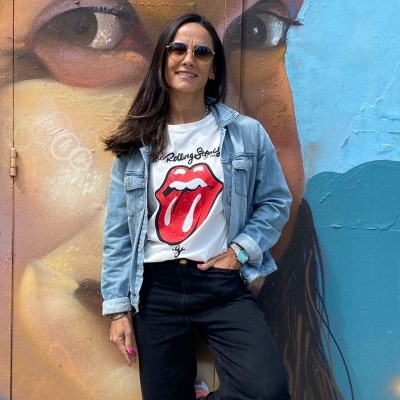Mulher com t-shirt Rolling Stones e casaco azul encostada a parede com graffiti de rosto colorido.