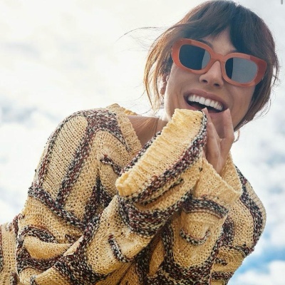 Pessoa com suéter de malha amarelo e castanho e óculos de sol castanhos sorrindo ao ar livre